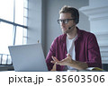 European man online tutor wearing glasses sitting at desk in headset and talking by video call 85603506