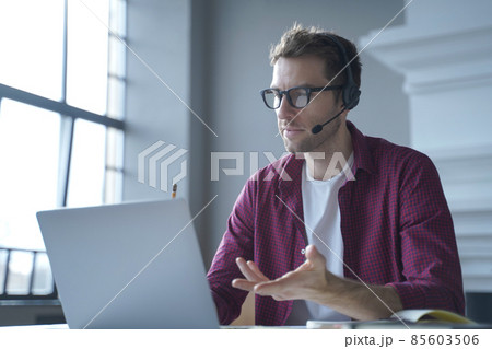 European man online tutor wearing glasses sitting at desk in headset and talking by video call 85603506