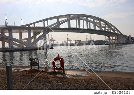 青空と雲と救命用の浮き輪のある陸と橋と海とクレーンやコンテナなどの港湾と霞むビル街の見える風景 85604135