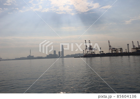 青空と雲と薄明光線と海面の少し光る海とクレーンの見える港湾と高層ビルのある街並みの風景 85604136