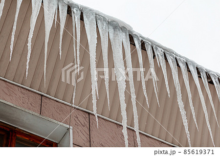 Large and long icicles hang from the roof of the building. Icicles create a danger for pedestrians. 85619371