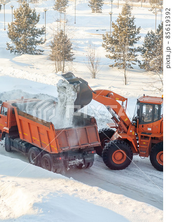 Big orange tractor cleans up snow from the road and loads it into the truck. Cleaning and cleaning of roads in the city from snow in winter. Snow removal after snowfall and blizzards. Big orange tractor cleans up snow from the road and loads it into the truck. Cleaning and cleaning of roads in the city from snow in winter. Snow removal after snowfall and blizzards. 85619392
