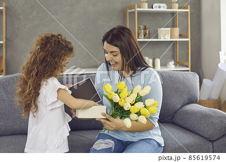 Lifestyle moment. Little daughter congratulates her loving mother on holiday by giving gifts and flowers. Happy young excited woman who is delighted with gift looks at him sitting at home on couch. 85619574