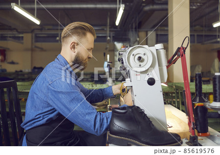 Profile portrait of shoe factory worker in workshop. Side view serious man sitting at table and using industrial sewing machine to make new black leather boots. Footwear manufacturing industry concept Profile portrait of shoe factory worker in workshop. Side view serious man sitting at table and using industrial sewing machine to make new black leather boots. Footwear manufacturing industry concept 85619576