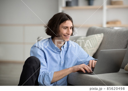 Blogging concept. Young guy using laptop, writing post to social network while sitting on floor near couch, free space 85620498