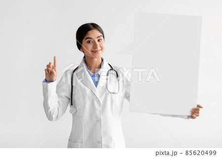Smiling young indian female doctor in uniform show finger up and hold big banner Smiling young indian female doctor in uniform show finger up and hold big banner 85620499