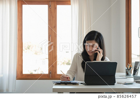 Happy business asian manageress working at her desk in the office taking a call on her mobile phone while writing notes on a notepad 85626457