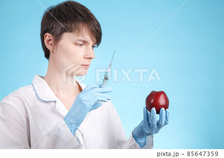 Portrait of a female doctor standing with an Apple and a syringe on blue as idea of antibiotics in vegetables and fruits 85647359