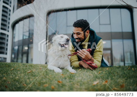 Happy young man lying on grass with his dog outdoors in town. 85649773