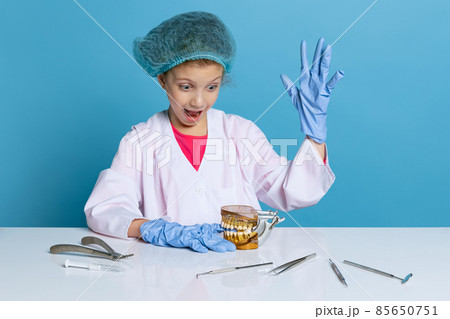 Emotional cute little girl, child in image of dentist doctor wearing white lab coat and gloves isolated on blue studio background 85650751