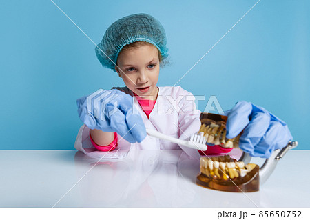 Emotional cute little girl, child in image of dentist doctor wearing white lab coat and gloves isolated on blue studio background Emotional cute little girl, child in image of dentist doctor wearing white lab coat and gloves isolated on blue studio background 85650752