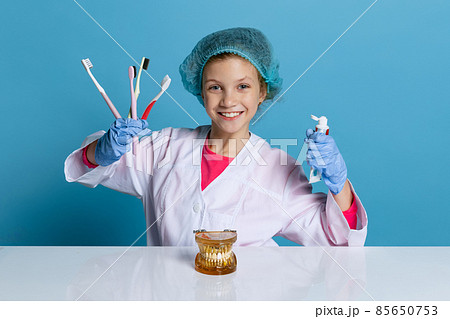 Emotional cute little girl, child in image of dentist doctor wearing white lab coat and gloves isolated on blue studio background 85650753