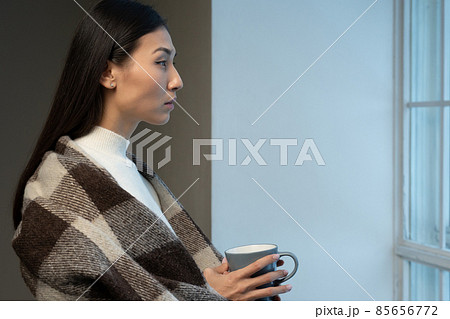 Sad Asian woman, covered with a blanket, drinks coffee or tea and looks out at the street from large modern windows Sad Asian woman, covered with a blanket, drinks coffee or tea and looks out at the street from large modern windows 85656772