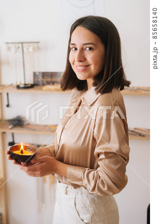 Vertical portrait of smiling young woman holding burning scented handmade candle in palms, looking at camera. 85658140