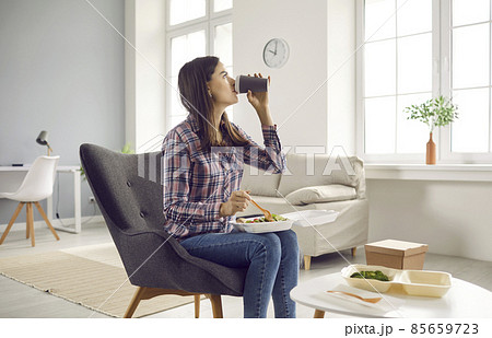 Woman having takeaway lunch at home. Beautiful young girl sitting in armchair in living room, drinking coffee and eating healthy vegetable salad from food container. Meal delivery service concept Woman having takeaway lunch at home. Beautiful young girl sitting in armchair in living room, drinking coffee and eating healthy vegetable salad from food container. Meal delivery service concept 85659723