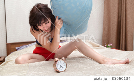 An angry woman in a rage hits an alarm clock with a pillow while sitting on the bed 85660184