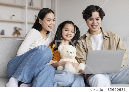 Cheerful young chinese male, female and teen daughter with toy watch video on laptop, searching, sits on sofa Cheerful young chinese male, female and teen daughter with toy watch video on laptop, searching, sits on sofa 85660621