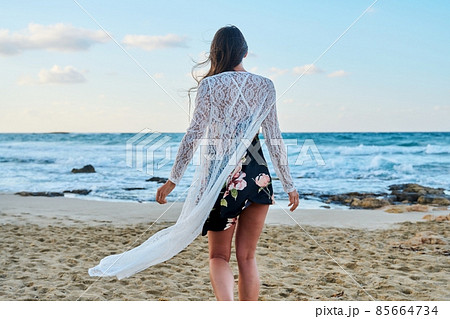Back view of young woman in fashionable romantic clothes walking along beach Back view of young woman in fashionable romantic clothes walking along beach 85664734