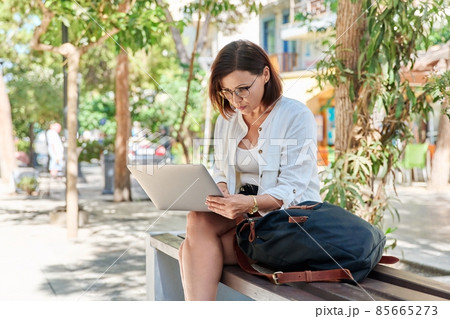 Middle-aged serious woman typing text on laptop outdoors Middle-aged serious woman typing text on laptop outdoors 85665273