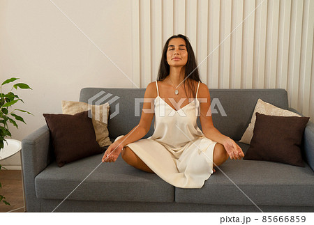 Young peacful woman sitting with eyes closed in lotus position on sofa at home, meditating in morning 85666859