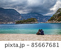 Popular Oludeniz beach on the background of the mountains in pre-thunderstorm weather. There are a couple of people sitting on the pebble shore. Oludeniz, Mugla, Turkey. 85667681