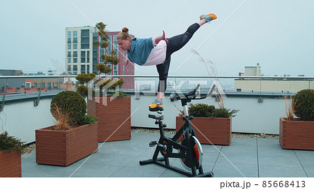 Athletic girl performing swallow acrobatic trick exercises on cycling stationary bike on house roof Athletic girl performing swallow acrobatic trick exercises on cycling stationary bike on house roof 85668413