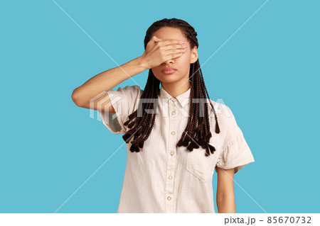 Young woman with black dreadlocks covering her eyes with palm, tries to hide from everybody, dontwant see someone face, wearing white shirt. Indoor studio shot isolated on blue background. 85670732
