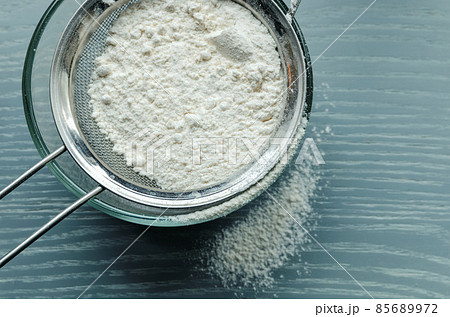 Wheat flour is sown through a sieve into a glass bowl through a metal sieve, on a wooden table. Wheat flour is sown through a sieve into a glass bowl through a metal sieve, on a wooden table. 85689972