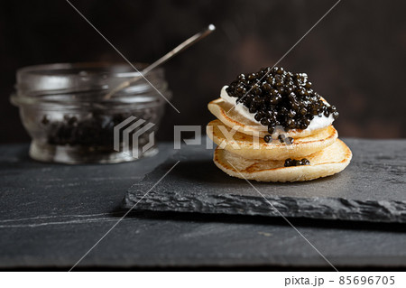 Black sturgeon caviar on small pancakes blinis with sour cream and a glass jar with caviar on a slate 85696705