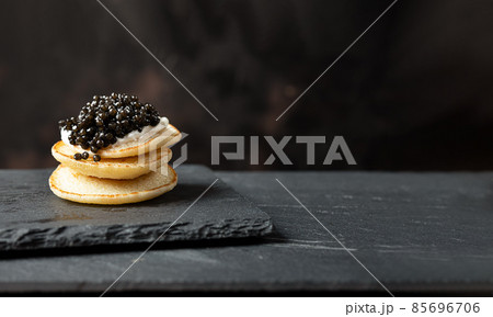 A stack of blini pancakes with black caviar and sour cream on a slate, copy space 85696706