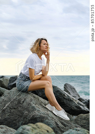 A young attractive Caucasian blonde woman of 35 years sits on the rocks of sea coast. Emerald-colored water, waves splashing on the stones. The woman meditates and relaxes. 85701755