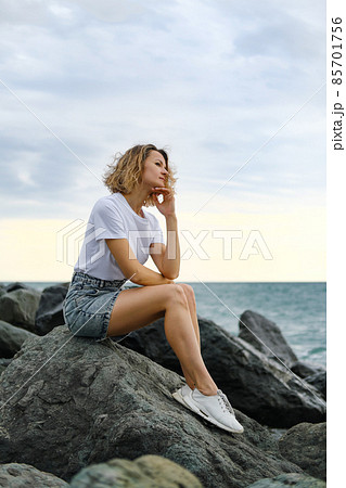 A young attractive Caucasian blonde woman of 35 years sits on the rocks of sea coast. Emerald-colored water, waves splashing on the stones. The woman meditates and relaxes. 85701756