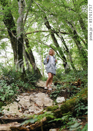 A young Caucasian woman walks along a forest mountain road. Casual blue denim clothes and backpack. Travel, healthy lifestyle and freedom. 85701757