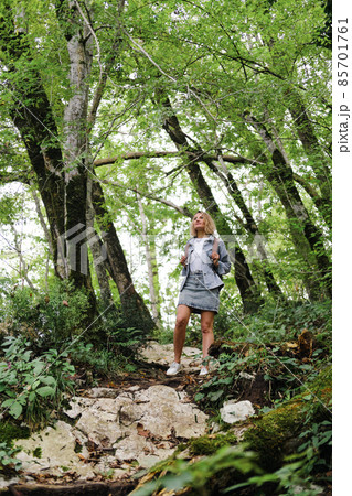 A young Caucasian woman walks along a forest mountain road. Casual blue denim clothes and backpack. Travel, healthy lifestyle and freedom. A young Caucasian woman walks along a forest mountain road. Casual blue denim clothes and backpack. Travel, healthy lifestyle and freedom. 85701761