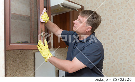 A tall adult man is cleaning the kitchen, wiping the cabinets 85702226