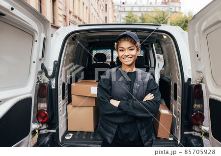 Portrait of a smiling delivery girl wearing a cap and uniform standing with crossed arms at van trunk Portrait of a smiling delivery girl wearing a cap and uniform standing with crossed arms at van trunk 85705928