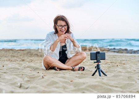 Middle aged woman sitting on beach with smartphone using video call Middle aged woman sitting on beach with smartphone using video call 85706894