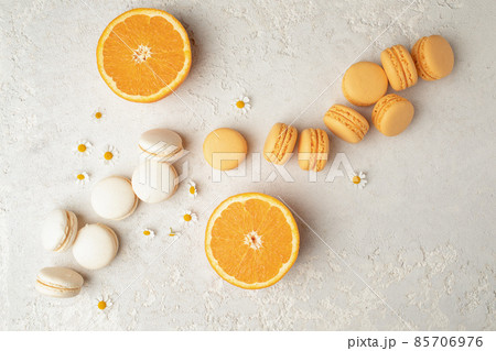 Orange and Vanilla French Macaroons with orange fruit filling on a creamy white clay background, decorated with small chamomiles and fresh sliced oranges 85706976