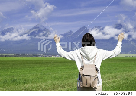 A woman tourist in a white hoodie and a backpack enjoys a beautiful view of the Tatra mountains while standing in a green field. Clouds float over mountain peaks, copy space 85708594