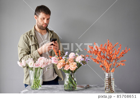 Male florist with a mobile phone, takes a photo to confirm the order. Accepts an online order for flower delivery. Responds to messages in social networks and online messengers. Male florist with a mobile phone, takes a photo to confirm the order. Accepts an online order for flower delivery. Responds to messages in social networks and online messengers. 85708651