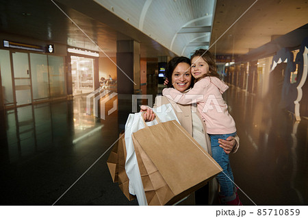 Happy mother holding daughter in arms, hugging her, smiling, looking at camera showing shopping bags standing in duty free area of departure international airport terminal, waiting for flight 85710859