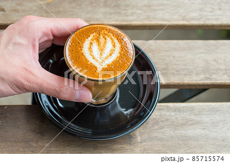 Close-up of human hand touching a cup of Piccolo latte on the wooden table. A Piccolo Latte is a ristretto shot topped with warm, silky milk served in a 100 ml glass demitasse (small latte glass). 85715574