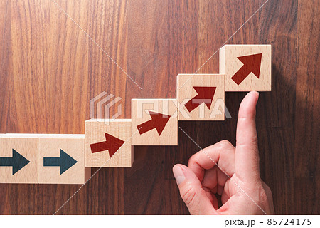 変化と成長のイメージ。矢印が描かれた積み木を指で押し上げる。木のテーブルの俯瞰。 85724175