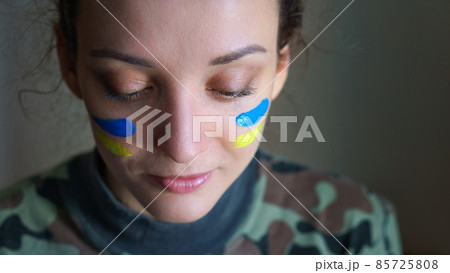 Indoor portrait of young girl with blue and yellow ukrainian flag on her cheek wearing military uniform, mandatory conscription in Ukraine, equality concepts 85725808
