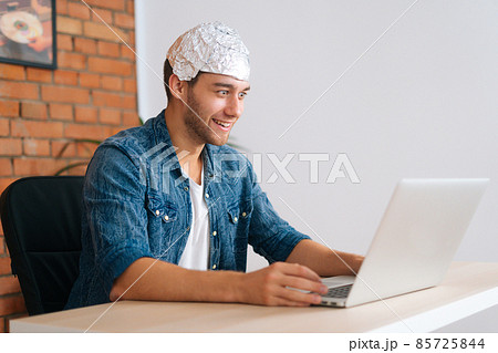 Medium shot of crazy male conspiracy theorist wearing aluminum foil protect brain watching shocked online video content using laptop computer. Medium shot of crazy male conspiracy theorist wearing aluminum foil protect brain watching shocked online video content using laptop computer. 85725844