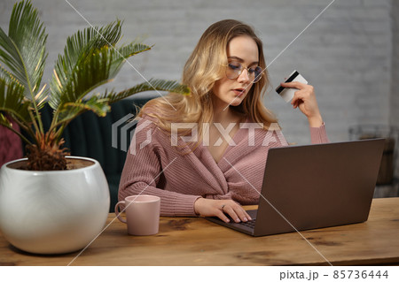 Female in pink cardigan, glasses. Working on laptop and showing white plastic card. Sitting at wooden table with pink cup, palm in pot on it. Close up 85736444