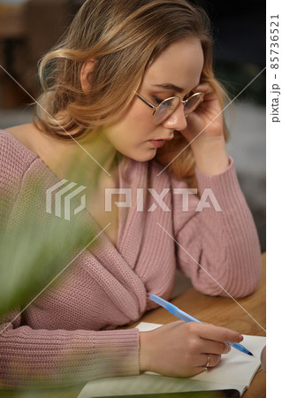 Lady in glasses, pink cardigan. Holding blue pen, sitting at wooden table with a tablet, notebook and green flower on it. Student, blogger. Close-up Lady in glasses, pink cardigan. Holding blue pen, sitting at wooden table with a tablet, notebook and green flower on it. Student, blogger. Close-up 85736521