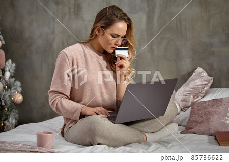 Blonde model blogger in glasses, casual clothing. Holding plastic card and working on her laptop, sitting on bed with book and cup. Close up Blonde model blogger in glasses, casual clothing. Holding plastic card and working on her laptop, sitting on bed with book and cup. Close up 85736622
