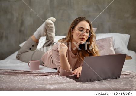 Blonde woman blogger in glasses, casual clothing. Holding plastic card, lying cross-legged on bed with laptop, book and cup. Work, education. Close-up 85736637