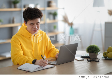 Asian man sitting at desk, using pc writing in notebook 85737708